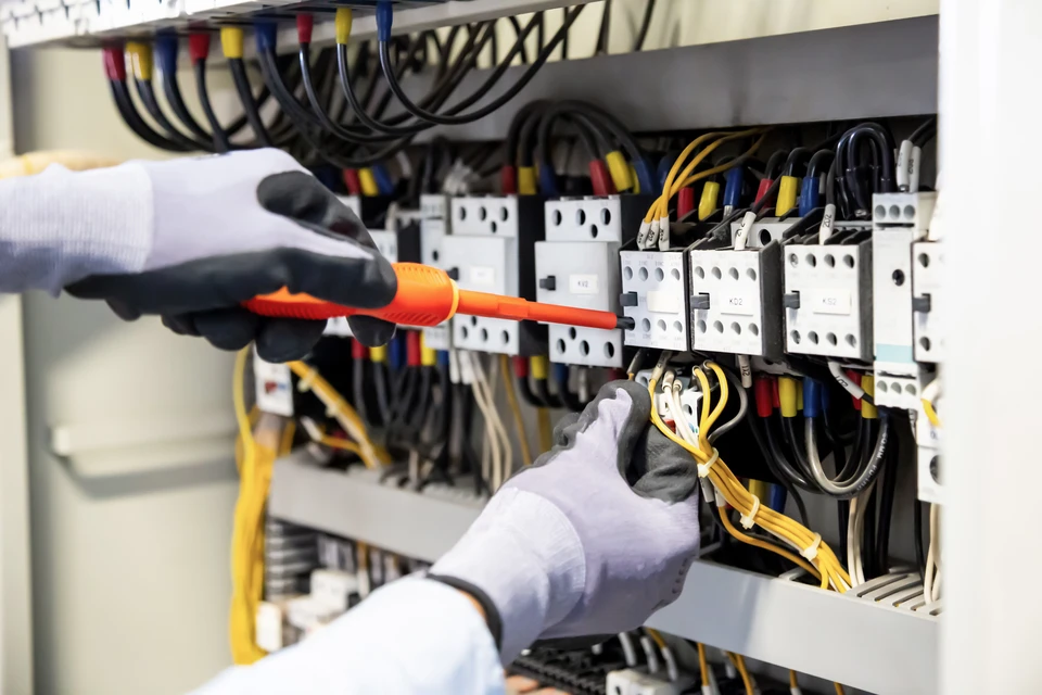 Un électricien en gants blancs travaille sur un tableau électrique, tenant un tournevis isolé orange
