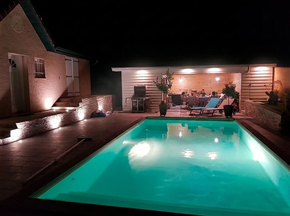 Soirée estivale en extérieur, groupe de personnes attablées sous un pool house ouvert en bois clair, éclairé par spots encastrés